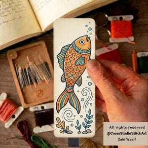 May include: A rectangular bookmark featuring a stylized orange fish with teal and yellow patterned scales, surrounded by small blue bubbles, swirling water currents, and aquatic plants on a cream-colored background with a smooth finish.