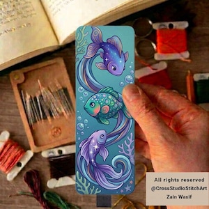 May include: A rectangular bookmark featuring a vertical illustration of three stylized fish swimming among bubbles and coral. The top and bottom fish are purple with star accents, while the middle fish is teal and green with scales.