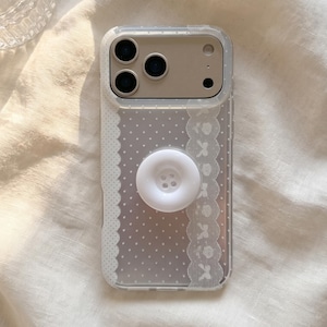 Op de afbeelding: Een doorzichtig smartphonehoesje met een wit stippenpatroon en een verticale kanten rand met een bloemendesign. Een ronde, witte, knoopvormige grip met vier gaatjes is bevestigd in het midden van de achterkant van het hoesje met een triple camera-uitsparing.
