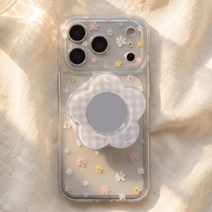 May include: A clear smartphone case featuring a pattern of small, multi-colored flowers in white, pink, yellow, and blue. Attached to the back is a light blue and white gingham-patterned, flower-shaped phone grip with a circular center.