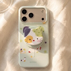 Op de afbeelding: Een lichtgroen smartphonehoesje met een ruitpatroon, voorzien van een uitsparing voor drie camera's en een ronde, uittrekbare grip in de vorm van een lapjeskat-gezicht. Het hoesje is versierd met kleine sterretjes, een sok en het woord KITTEN in zwart.