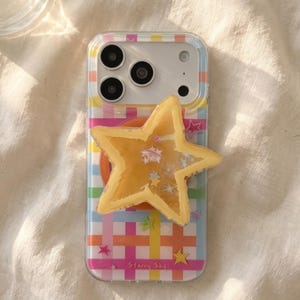 May include: A clear smartphone case featuring a colorful plaid pattern of pink, orange, yellow, green, and blue stripes. A large, yellow, star-shaped phone grip is attached to the center, containing small star-shaped glitter. The text Starry Sky is printed at the bottom.