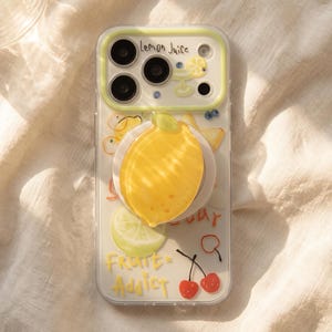 May include: A clear smartphone case featuring a yellow lemon-shaped pop-out grip, a light yellow camera frame, and various fruit illustrations including lemons, limes, and cherries. The text "Lemon Juice" and "Fruit Addict" is printed on the case.