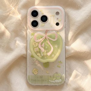 May include: A clear smartphone case featuring a light green apple-shaped phone grip with a pink bow. The case is decorated with small green apple illustrations, stars, and a checkered pattern at the bottom with the text A for Apple, That we share so the case.