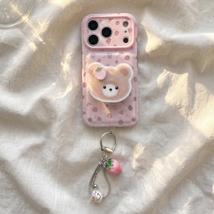 May include: A light pink smartphone case featuring a repeating strawberry pattern and a raised, three-dimensional bear-shaped grip on the back. A silver-toned metal keychain with a small pink strawberry charm and a white bead is attached below.