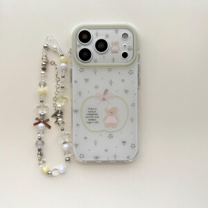 May include: A clear phone case featuring a printed pattern of small stars, bows, and a central apple-shaped graphic containing a teddy bear and the text There is always unexpected warmth and endless hope in life, paired with a beaded phone strap.