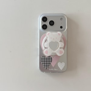 May include: A clear smartphone case featuring a white bear-shaped phone grip with a heart-shaped centre. The case is decorated with a large pink heart, a black and white gingham heart, a small white polka dot heart, and the text Little Bear.