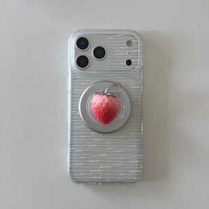 May include: A clear smartphone case featuring thin horizontal white lines and small white text patterns. Attached to the center is a circular magnetic mount topped with a three-dimensional, textured pink and red strawberry charm with a silver stem.
