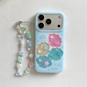 May include: A light blue smartphone case featuring four iridescent shell-shaped cutouts filled with small star charms, accompanied by a beaded phone strap with clear, green, blue, and pink beads shaped like stars, hearts, and seashells.