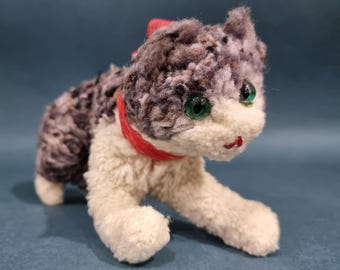 Puede incluir: Un juguete de peluche con forma de gato con un pelaje texturizado de color gris y blanco moteado, patas delanteras de color crema y un collar rojo brillante. El gato tiene ojos de plástico verde y una pequeña nariz roja bordada.