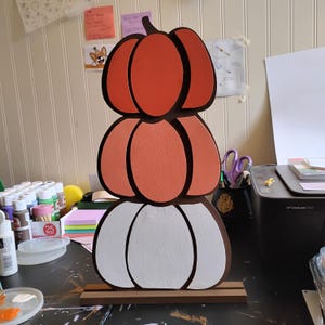 May include: A vertical stack of three wooden pumpkin cutouts mounted on a rectangular base. The top pumpkin is dark orange, the middle is a lighter coral orange, and the bottom is white. Each pumpkin shape is outlined with a dark brown border.