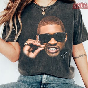 May include: A charcoal grey vintage-wash t-shirt featuring a large graphic print of a person wearing dark sunglasses and gold teeth. The print includes the signature Usher in white script. The shirt has short sleeves and a crew neckline.