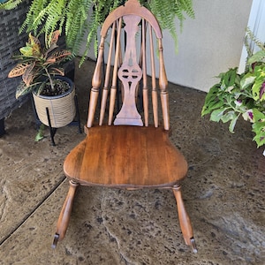 May include: A wooden rocking chair with a medium brown finish, featuring a high backrest with vertical spindles and a central decorative splat with a cutout design. The chair has a smooth, contoured seat and two curved rockers at the base.