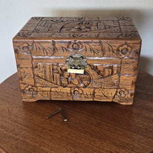 May include: A rectangular wooden chest with intricate hand-carved floral and landscape patterns on the lid and front panels. The box features a brass-colored metal latch mechanism at the center and includes a small, separate metal key component.