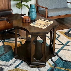 May include: A hexagonal dark wood side table with a lower shelf and decorative cutouts on the legs. On the tabletop sits a teal ceramic vase, a stack of round wooden coasters, and a magazine with the word domus printed on the cover.