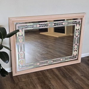 May include: A rectangular wall mirror with a light pink wooden frame. The glass features a decorative border with a geometric pattern in shades of sage green, soft pink, and cream, incorporating stylized floral and leaf motifs along the edges.