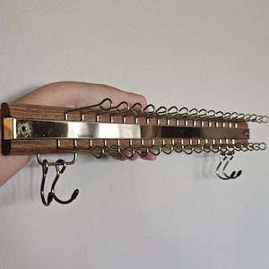 May include: A wall-mounted tie rack featuring a horizontal dark wood base with a central polished brass strip. The rack includes multiple gold-toned wire loops extending from the top and bottom, along with two small metal hooks with black tips.