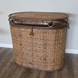 May include: An oval-shaped brown wicker hamper with a woven lid. The hamper features a dark brown leather-like strap with gold-toned metal rings and a central latch mechanism. The structure is reinforced with a solid brown rim at the top and base.