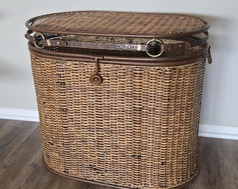 May include: An oval-shaped brown wicker hamper with a woven lid. The hamper features a dark brown leather-like strap with gold-toned metal rings and a central latch mechanism. The structure is reinforced with a solid brown rim at the top and base.