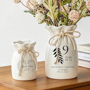 May include: Two white ceramic vases with ruffled rims and tied rope accents. The larger vase features a black leaf graphic, the number 9, and the text DELILAH & CAROLINE 9 YEARS 09.11.2027. The smaller vase displays a floral heart design with text.