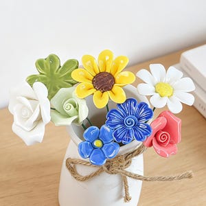May include: A white ceramic vase tied with a jute twine bow, containing a bouquet of colorful ceramic flowers including a yellow sunflower, white and pink roses, a white daisy, a green clover, and blue blossoms with yellow centers.