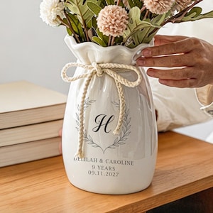 May include: A white ceramic vase with a ruffled top edge and a beige rope tied in a bow around the neck. The front features a black printed laurel wreath surrounding the letter H, with the text DELILAH & CAROLINE, 9 YEARS, and 09.11.2027 below.