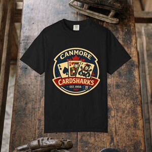 May include: A black short-sleeve t-shirt featuring a circular graphic with the text CANMORE CARDSHARKS EST. 1956. The design includes playing cards, a king, a jester, a red maple leaf, and a small flag emblem in red, blue, and cream colors.