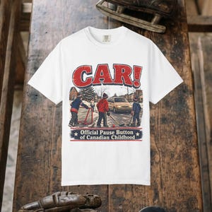 May include: A white short-sleeve t-shirt featuring a graphic of children playing street hockey in a neighborhood. The text reads CAR! at the top and Official Pause Button of Canadian Childhood at the bottom in red, white, and blue lettering.