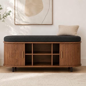 May include: A rectangular wooden storage bench with a dark charcoal gray upholstered seat cushion. The base features vertical slatted wood paneling, two side cabinets with thin gold handles, and an open center section with four storage cubbies.
