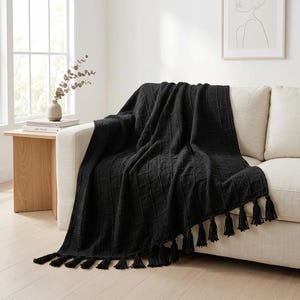 May include: A black throw blanket with a textured grid pattern and fringe tassels along the bottom edge. The blanket is draped over the arm and seat of a light-colored sofa, showcasing its woven fabric and decorative knotted details.