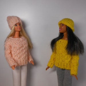 Può includere: Due bambole che indossano maglioni e cappelli di lana. Una bambola ha un maglione e un cappello rosa, l'altra ha un maglione e un cappello giallo.