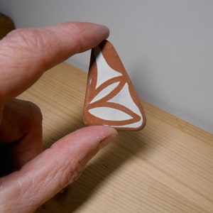 Könnte beinhalten: Eine dreieckige Keramikfliese mit einem weißen Design aus drei sich überlappenden Tropfenformen auf braunem Hintergrund.