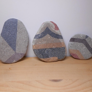Può includere: Tre pietre decorative con motivi geometrici in tonalità di grigio, rosa e marrone. Le pietre sono disposte in fila su una superficie di legno.