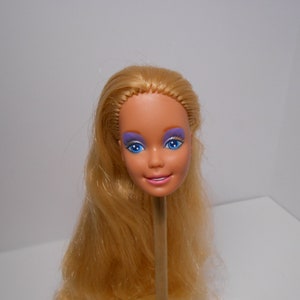 Könnte beinhalten: Ein blonder Barbie-Puppenkopf mit blauen Augen und lila Lidschatten. Der Kopf sitzt auf einem Holzstab.