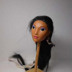 Può includere: Una testa di bambola con lunghi capelli neri, pelle marrone e ombretto blu e viola. La testa è montata su un  perno di legno.