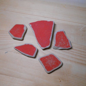 Peut inclure: Cinq carreaux de céramique rouges de formes et de tailles différentes, avec une surface rugueuse et texturée. Les carreaux sont disposés en grappe sur une surface en bois clair.