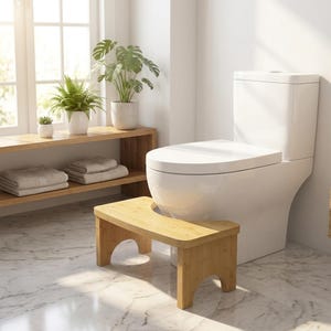 May include: A light-coloured bamboo toilet stool with a curved cutout design, positioned in front of a white ceramic toilet. The stool features a natural wood grain finish and four sturdy legs, providing a functional step for bathroom use.