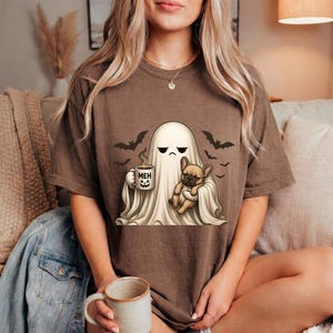 Könnte beinhalten: Ein braunes Kurzarm-T-Shirt mit einer Grafik eines weißen Geistes mit mürrischem Gesichtsausdruck, der eine Tasse mit der Aufschrift MEH und einem Kürbisgesicht hält. Der Geist hält eine beige französische Bulldogge, umgeben von schwarzen Fledermäusen.