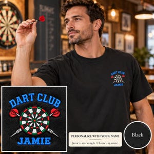 May include: A black t-shirt featuring an embroidered design on the left chest. The design includes a circular dartboard with two crossed darts and the text DART CLUB in blue above and the name JAMIE in blue below the dartboard graphic.