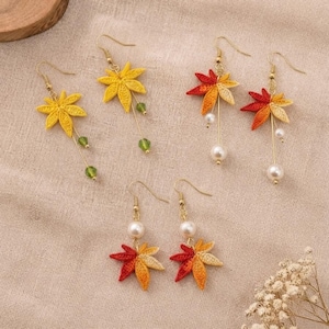 May include: Three pairs of drop earrings featuring embroidered maple leaf motifs. One pair is yellow with green beads, while two pairs feature red, orange, and yellow leaves with white faux pearl accents. All earrings have gold-toned metal hooks.
