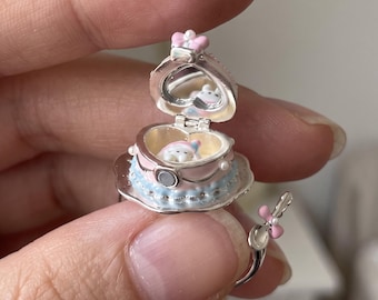 Könnte beinhalten: Ein silberfarbener Ring mit einem kleinen, herzförmigen Medaillon, das sich öffnen lässt und eine winzige, modellierte Figur im Inneren zeigt. Das Medaillon ist mit pastellrosa und blauen Emaille-Details, einem kleinen Spiegel und einer rosa Schleife verziert.