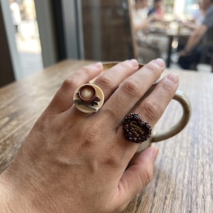 May include: Two rings worn on fingers. One ring features a miniature cream-colored saucer with a brown coffee cup, a heart-shaped foam design, and a small spoon. The other ring is heart-shaped, dark brown, textured with coffee bean shapes, and displays the text Coffee Time.