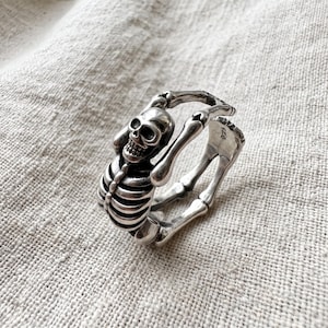 Op de afbeelding: Een zilverkleurige ring met een gedetailleerd skeletontwerp dat om de vinger wikkelt. De ringband is gevormd als menselijke botten, met de schedel aan de bovenkant en de ribbenkast aan de voorzijde. De binnenzijde is gestempeld met S925.
