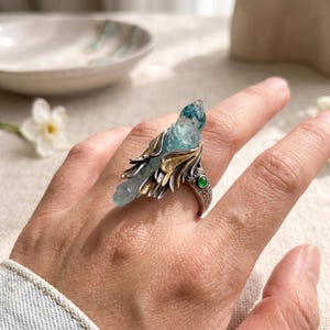 Puede incluir: Un anillo con una punta de cristal alargada de color azul claro, engastada en un marco de metal plateado y dorado con forma de alas. La banda tiene un acabado texturizado y está decorada con una pequeña piedra redonda de color verde vivo.