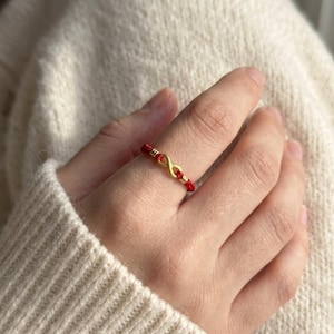 May include: A ring featuring a thin red cord band accented with a central gold-toned infinity symbol. The infinity charm is flanked by small gold-toned cylindrical beads on either side, creating a continuous loop design around the finger.