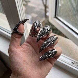 May include: A set of five dark metallic claw rings worn on the fingers. Each ring features a pointed, tapered design covered in small, reflective dark crystals or studs, extending over the fingernail area with a smooth metal base.