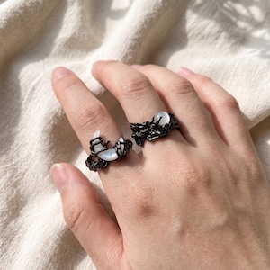 Könnte beinhalten: Zwei Ringe aus schwarzem Metall mit texturierten, organisch wirkenden Bändern. Ein Ring ist mit einem weißen, halbmondförmigen Stein besetzt, während der andere einen kleinen, runden, weißen Stein in der dunklen, unebenen Metallstruktur aufweist.