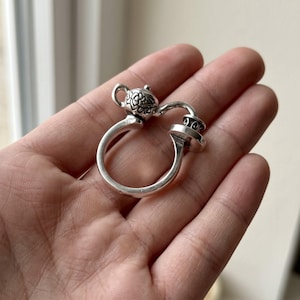 Puede incluir: Un anillo de plata con un diseño de tetera en miniatura en la banda. La tetera tiene un patrón floral en relieve en su cuerpo y un pico curvo que se extiende hacia una pequeña plataforma circular unida al lado opuesto de la banda del anillo.
