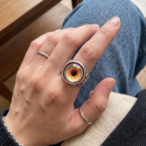 May include: A silver ring featuring a prominent, circular glass cabochon with a realistic, detailed eye design. The eye has a black pupil, a vibrant orange and yellow iris, and a reddish outer rim. The metal band displays intricate, embossed patterns.