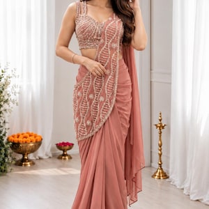 May include: A dusty rose saree featuring a sleeveless blouse with intricate silver beadwork and sequin embroidery in a wave pattern. The draped skirt includes a matching embellished panel and a plain flowing fabric, finished with a delicate beaded border.